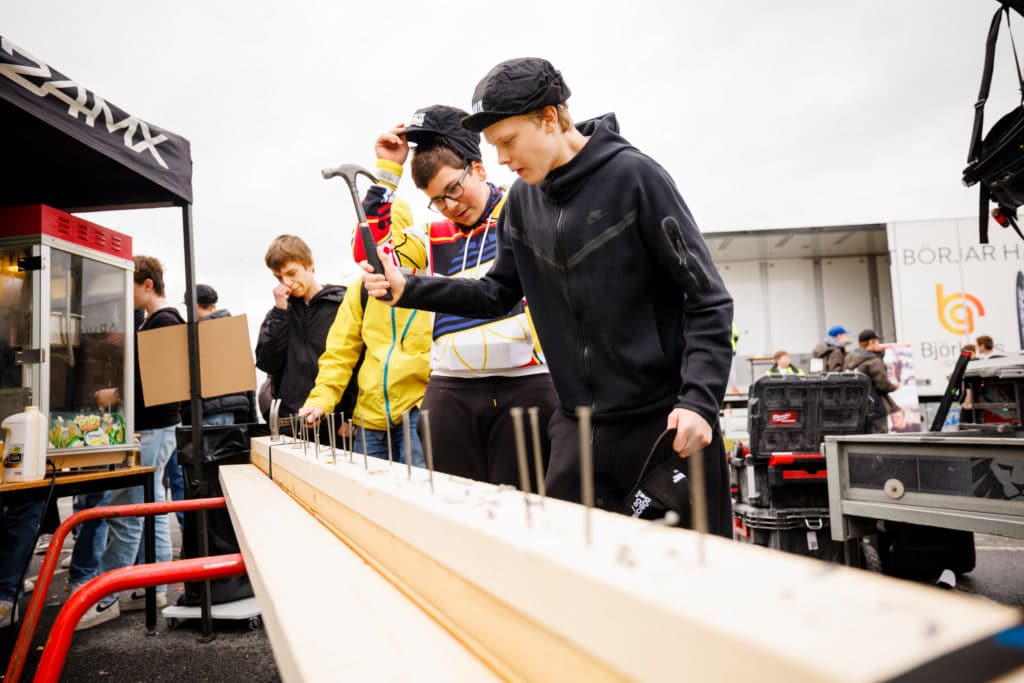 Three young boys are hammering nails on a plank outside.