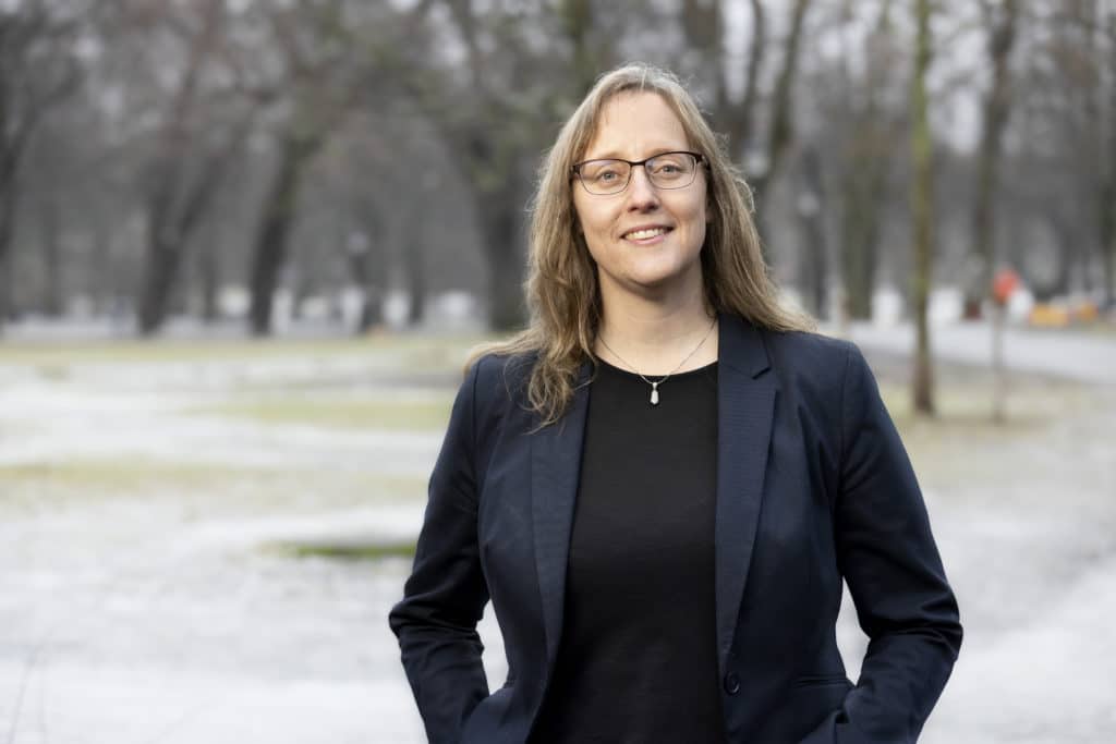 Woman standing with her hands in her pockets. She wears a jacket, glasses and looks happy. In the background are trees. 