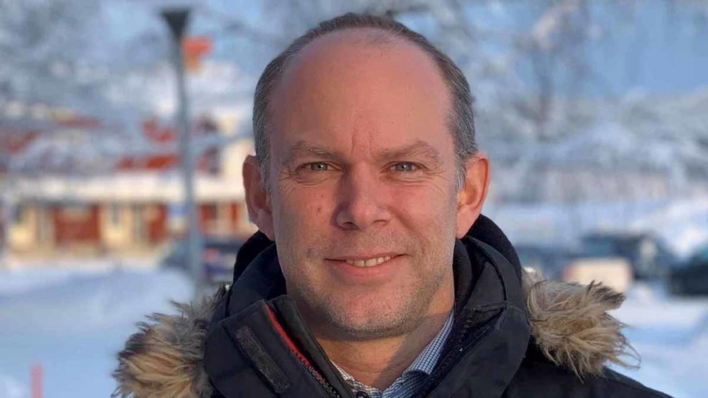 A man looks into the camera and looks happy. The man is wearing a warm jacket and appears to be standing outside during the winter. There is snow in the background. 