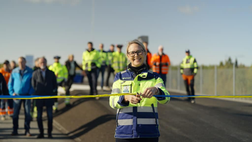 En kvinna i reflexjacka klipper ett band vid en invigning. I bakgrunden står en grupp personer i olika färgglada arbetskläder. Himlen är klarblå.