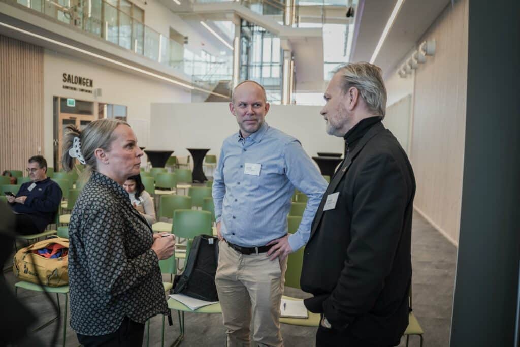 A general view of a modern and bright foyer where a group of people mingle and converse. The decor is contemporary with comfortable seating and green plants. A window section lets in daylight and provides a view of the outdoor area.