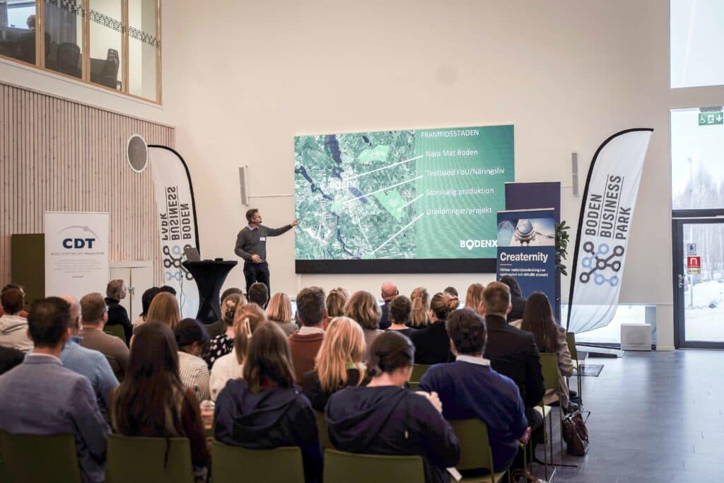 En talare presenterar framför en stor skärm med en satellitbild och texten "Framtidstaden" vid ett evenemang. Publiken, sittandes i en föreläsningssal, lyssnar uppmärksamt. Evenemanget verkar vara relaterat till lokala initiativ i Boden, såsom närproducerad mat och storskalig produktion. Banners för Boden Business Park och CDT, tillsammans med Bodenxt och Creaternity logotyper, tyder på ett fokus på innovation och näringslivsutveckling.