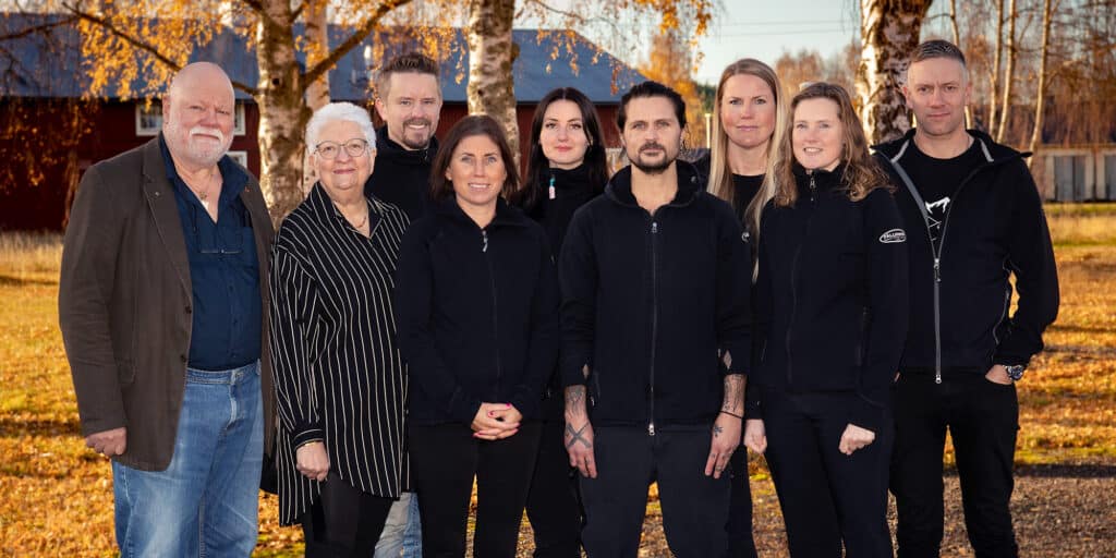 A group of eight adults stand in front of an autumnal setting of bare trees and a traditional red wooden building. The group is smiling and dressed in casual and work clothes, suggesting a relaxed and possibly professional environment.