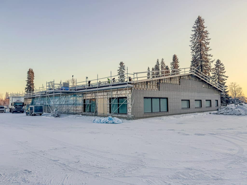 Ett vinterlandskap av en byggarbetsplats med en enplansbyggnad delvis täckt av byggnadsställningar. Snö täcker marken, och några barrträd står i bakgrunden, badade i det mjuka ljuset från en nedgående eller uppstigande sol.