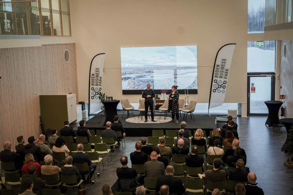 En man talar till en publik i en konferenssal. På bakgrunden visas Polar Structures logotyp och en bild av ett vinterlandskap, med texten "BODEN BUSINESS PARK".