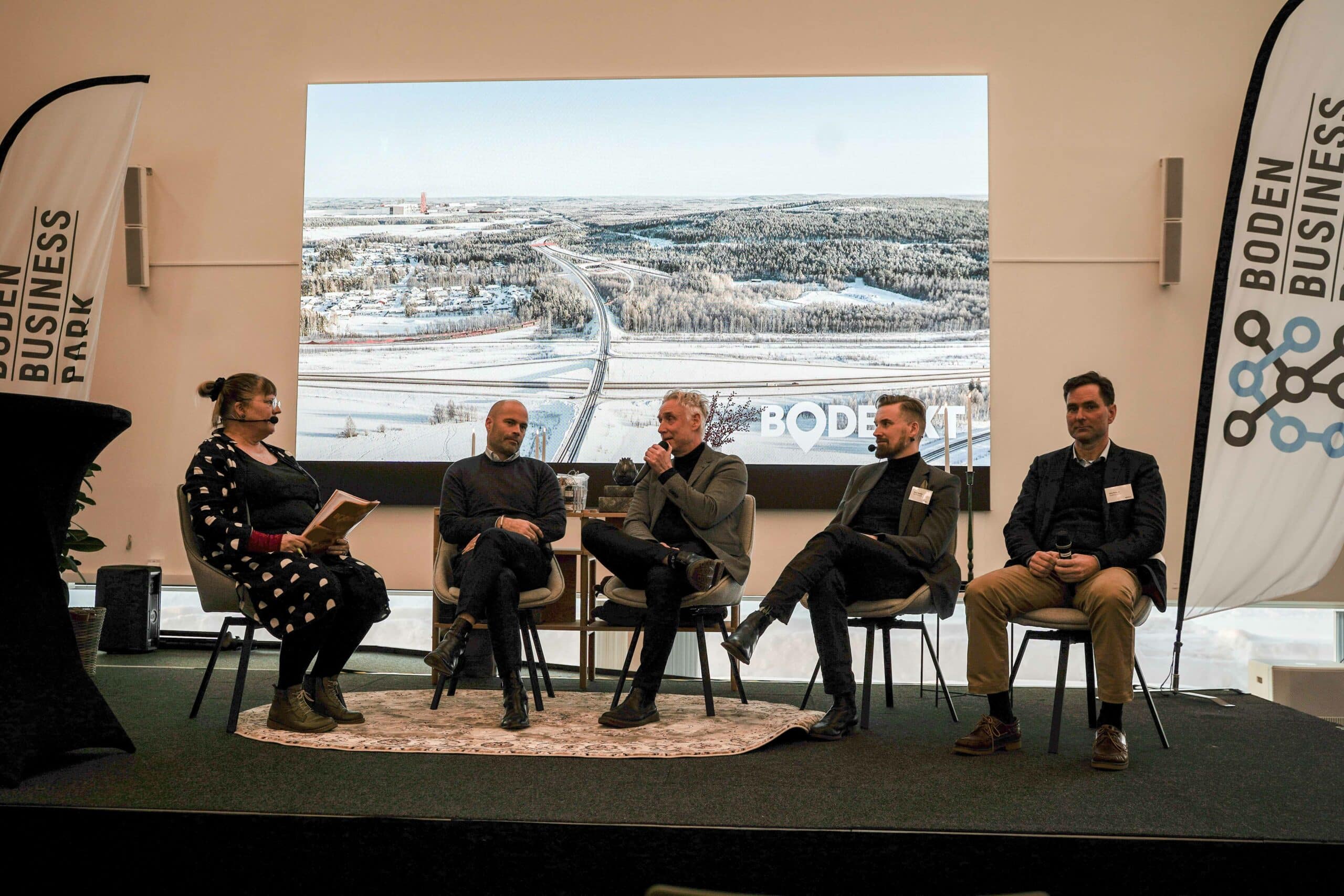 Bild 1: En panel med fyra deltagare och en moderator i en konferensmiljö. Bakom dem syns en stor skärm med en bild av ett snöigt landskap och texten "BODEN BUSINESS PARK".