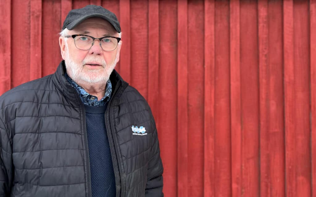 A picture of the same older man standing in front of a red wall. The man is wearing a black jacket and a cap, and he looks directly at the camera with a serious but friendly expression. The man has a logo on his jacket that appears to be for an organization or company.