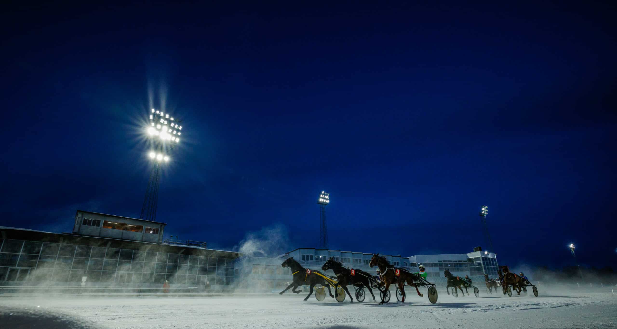 En bredvinklad bild som fångar ett travlopp under pågång på natten, med flera kuskar och travhästar som tävlar, och en snömoln som hästarna virvlar upp, under de starka ljusen från travanläggningen.