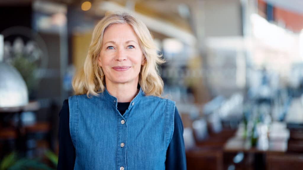 Sara Öhrvall with blonde hair in a denim vest and black sweater smiles at the camera. She is standing indoors with a blurred background suggesting that it is a café or a restaurant.