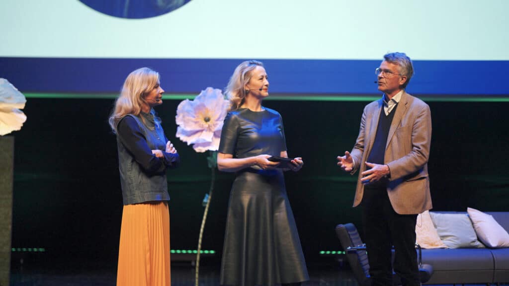 Three people on a stage take part in a discussion. A woman in a black dress stands in the center holding a tablet, while talking to a man in a beige jacket and a woman in an orange skirt and denim vest. Flowers are visible in the background of the scene.