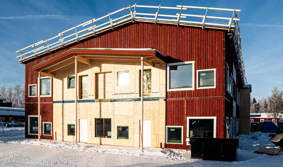 A two-story building under construction with a red-painted wooden facade and exposed wood paneling on the front. The building is surrounded by snow and scaffolding is visible on the roof under a clear blue sky.