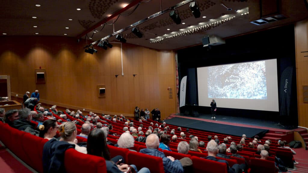 Half-filled auditorium at Folkets Hus in Boden during a Q&amp;A session with Stegra. On stage, Henrik Henriksson, CEO of Stegra, stands in front of a large screen with the company's logo. 