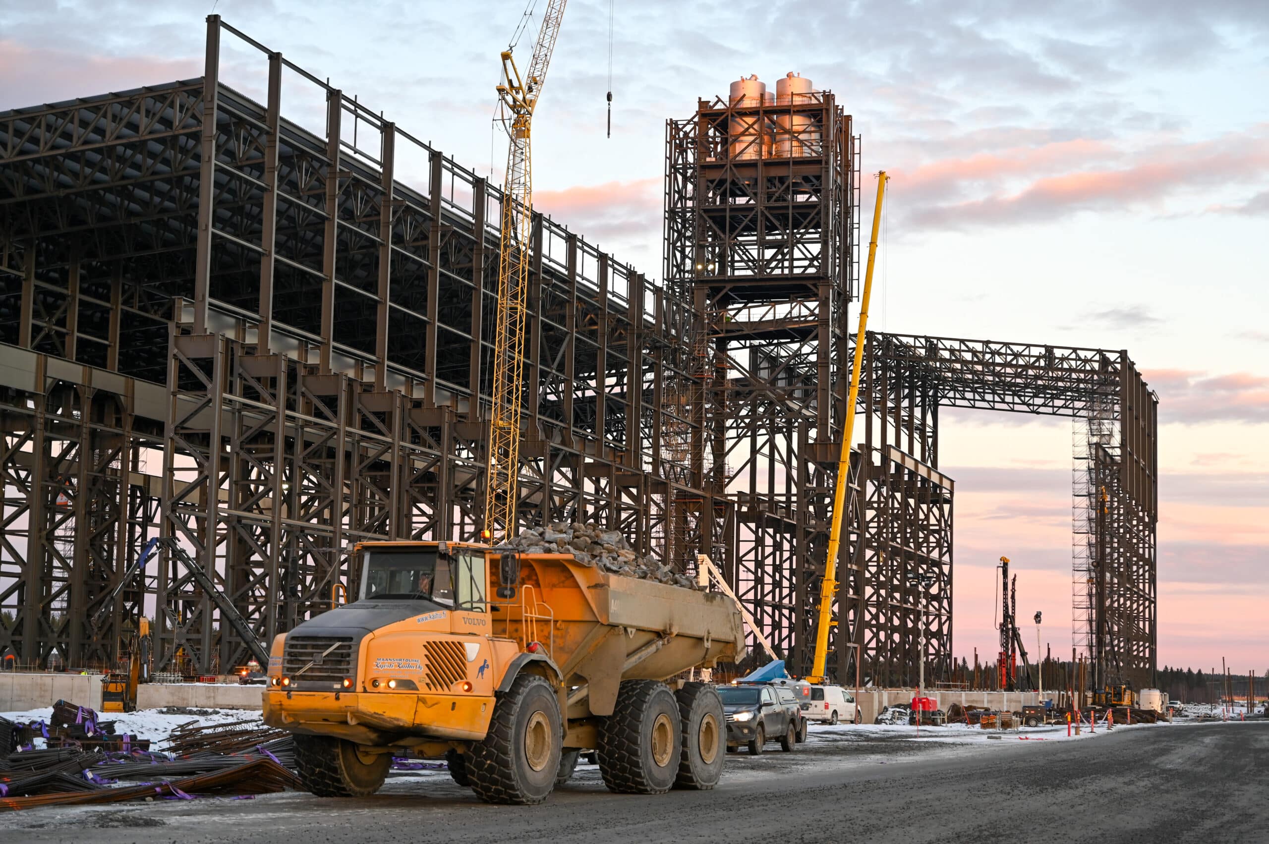 En industriell byggarbetsplats i gryning eller skymning med en stor stålstomme under uppbyggnad. En gul dumperlastbil lastad med sten syns i förgrunden, och flera kranar och arbetande fordon är utspridda på platsen. Himlen har rosa och blå toner, och marken är täckt av grus och fläckar av snö.