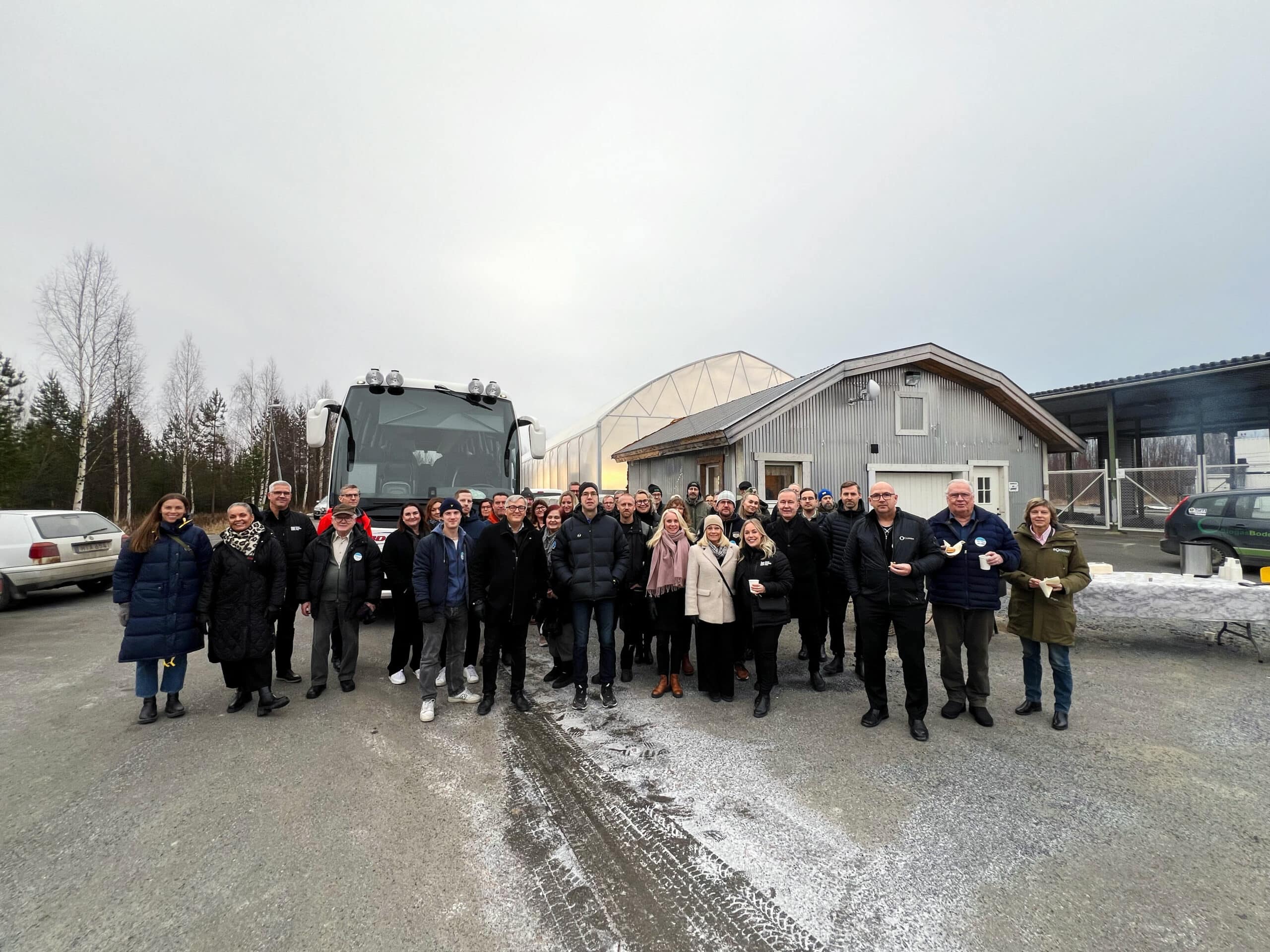 Gruppbild med ett stort antal människor stående framför en buss utomhus. De är klädda för kallt väder, och i bakgrunden syns en byggnad och vinterlandskap.