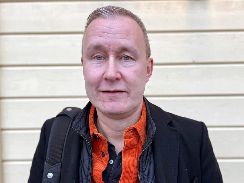 Portrait of a man with short hair, dressed in a black jacket and an orange shirt, photographed in front of a light wooden wall.