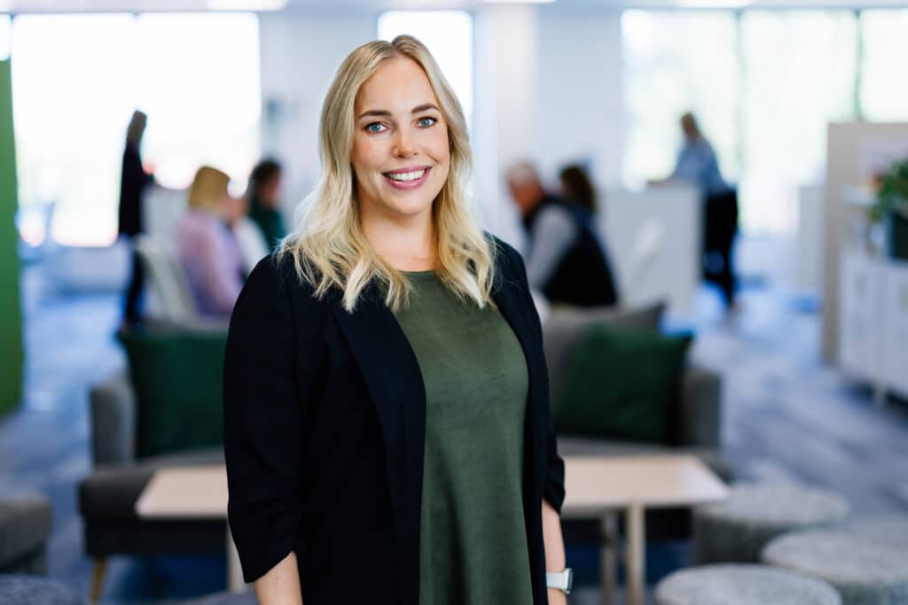 Portrait of a smiling woman with blonde hair, dressed in a black jacket and a green top. She stands in a modern office environment with a blurred background showing people at work. 