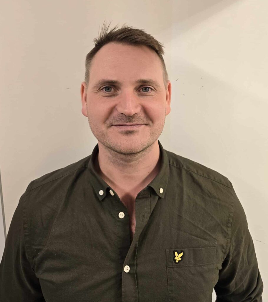 Portrait shot of a man with short brown hair, wearing a dark green shirt with a yellow logo on the breast pocket. He stands in front of a light background and looks into the camera with a neutral smile. 