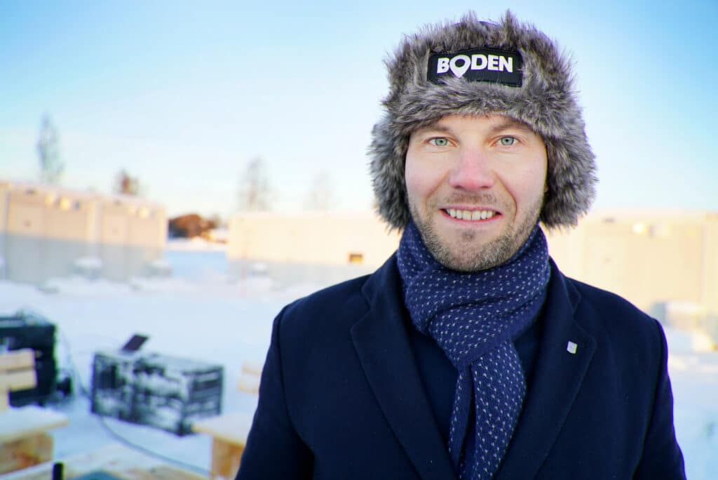 A man dressed in winter clothes stands outside in a snowy landscape. He is wearing a fur hat with the word 'Boden' on it, a dark jacket and a spotted scarf. The background shows buildings and trees in cold and sunny weather.  