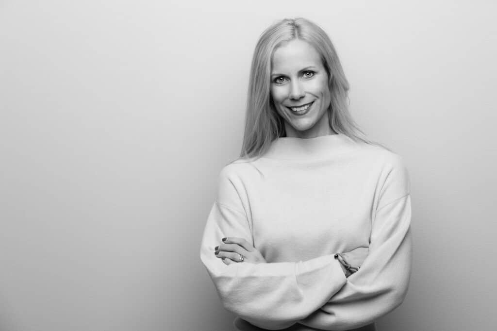 Black and white portrait of a smiling woman with long, fair hair. She is standing with her arms crossed in front of a light background and wearing a simple, stylish sweater. 