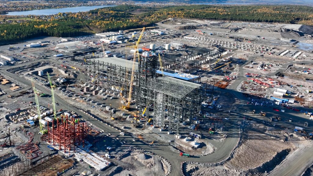 Aerial view of a large construction site with several steel structures under construction, cranes and surrounding forest landscape in autumn colors.