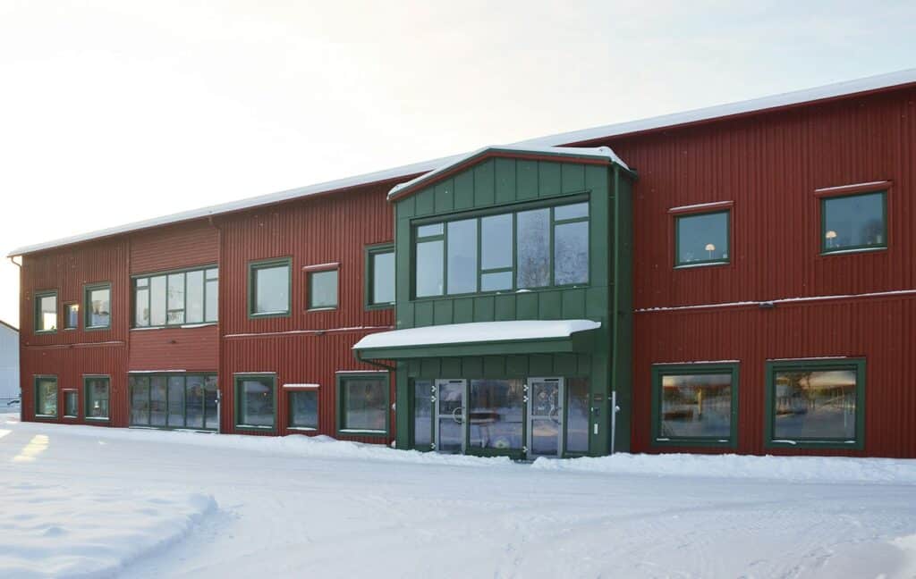 A two-storey building with a red wooden façade and green details around the windows and entrance, covered with snow.