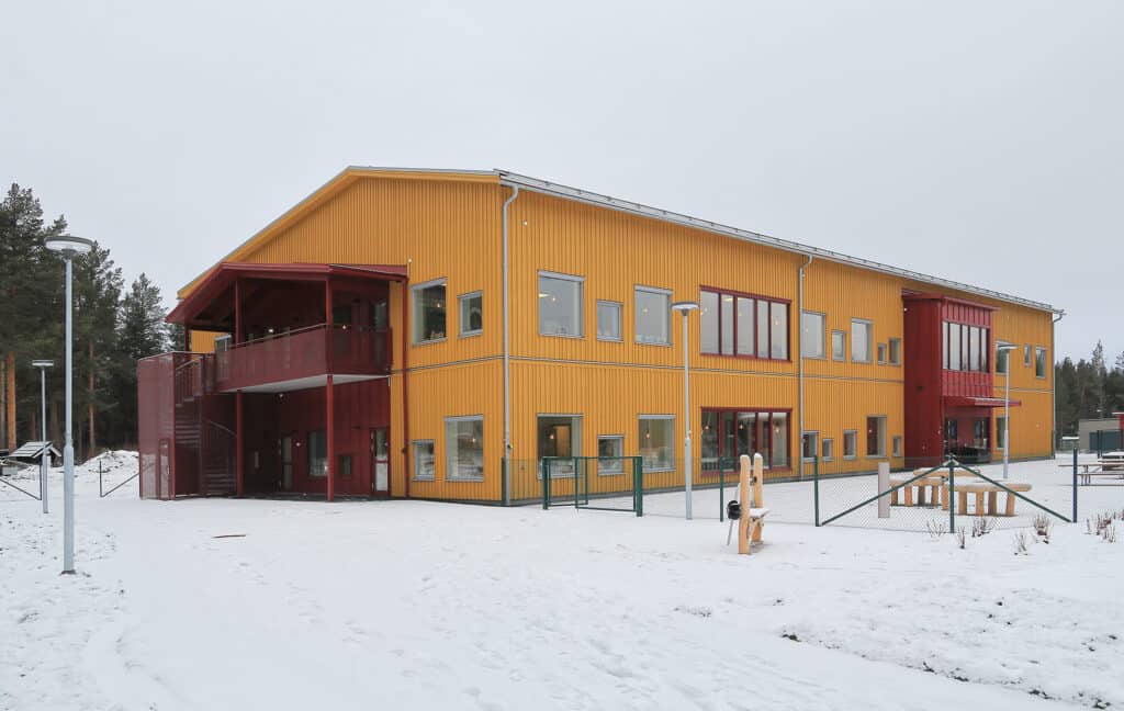 A school building in yellow and red, surrounded by snow, with a playground and gate in the foreground.