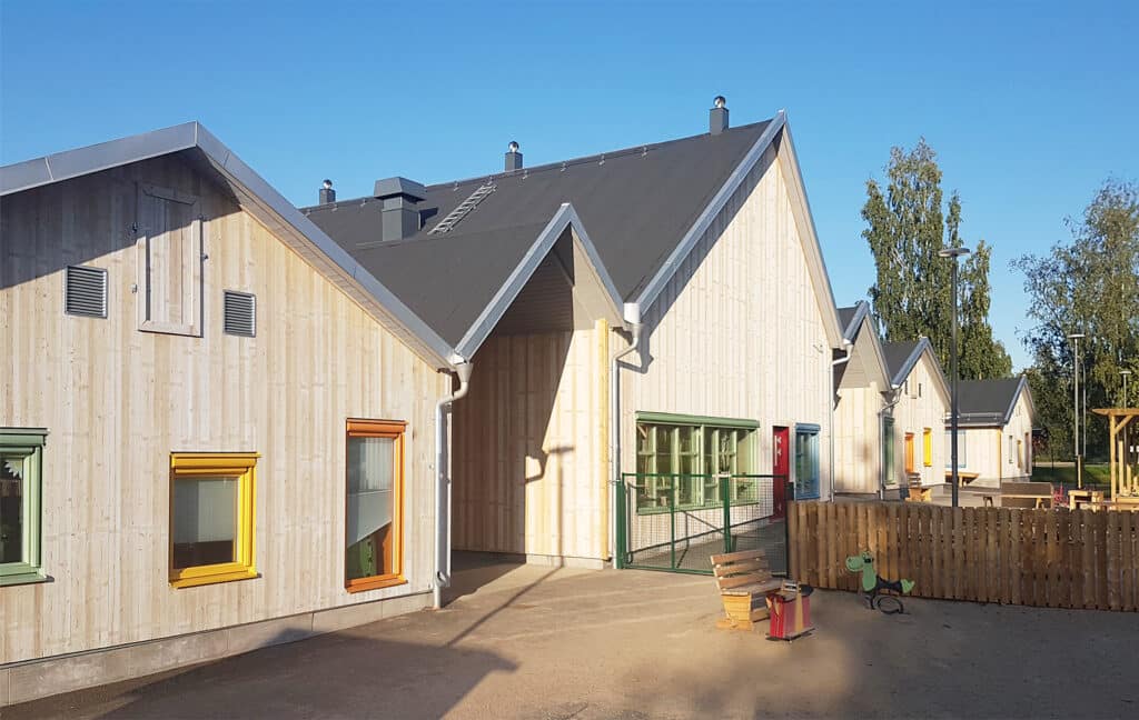 A modern preschool with light wood paneling and colorful window frames, with a small playground surrounded by wooden fences.